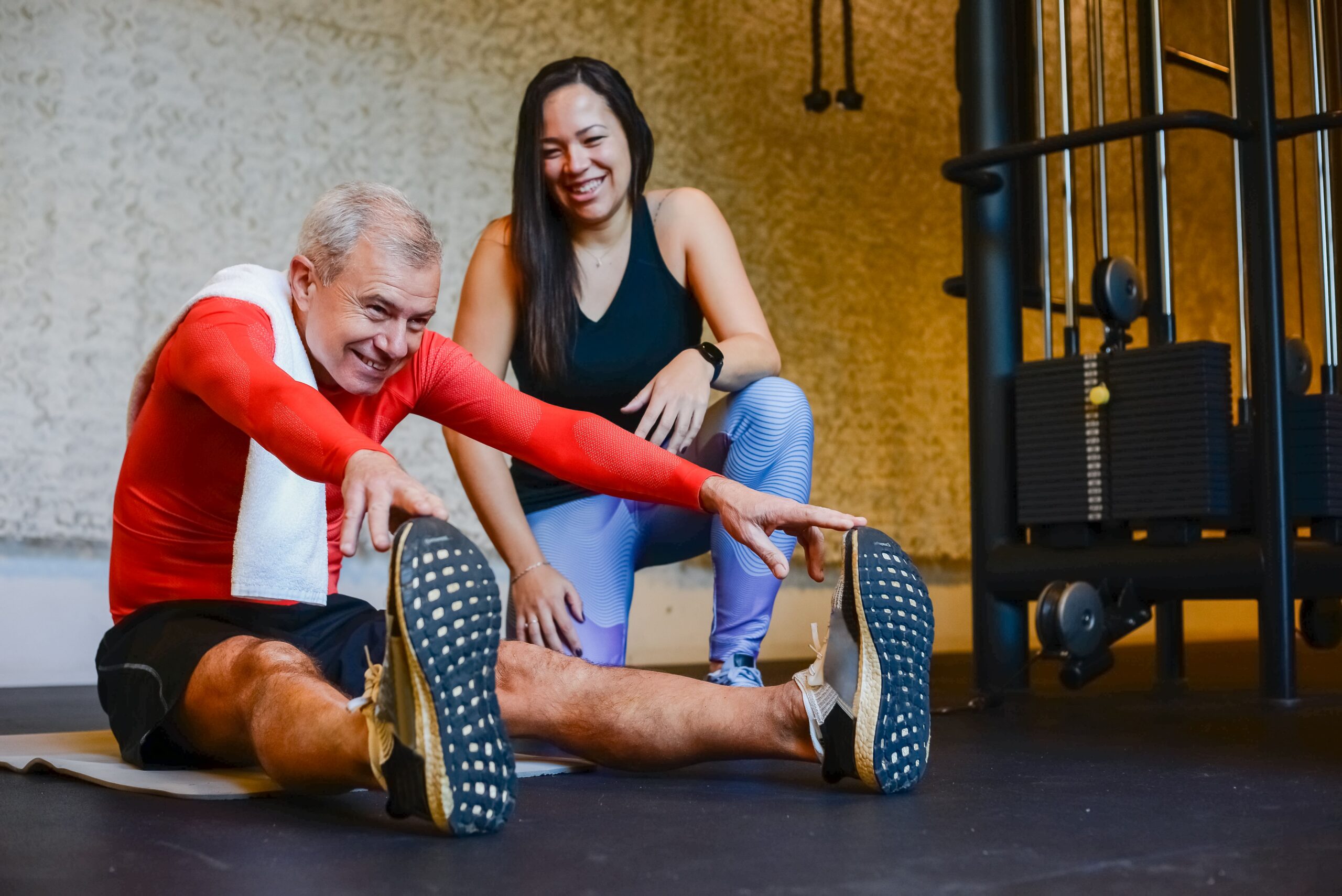 Elderly man in a gym stretching with a fitness coach, promoting a healthy lifestyle.